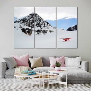May include: A red airplane sits on a snowy mountaintop with a blue sky and white clouds in the background.