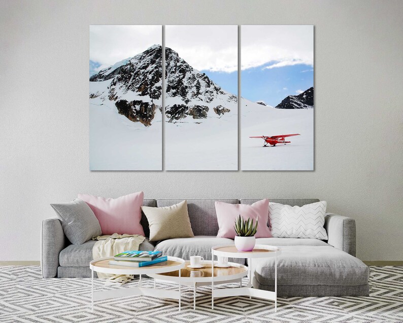 May include: A red airplane sits on a snowy mountaintop with a blue sky and white clouds in the background.