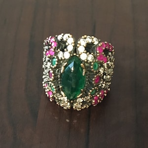 May include: A silver ring with a large green gemstone surrounded by smaller white and pink gemstones.