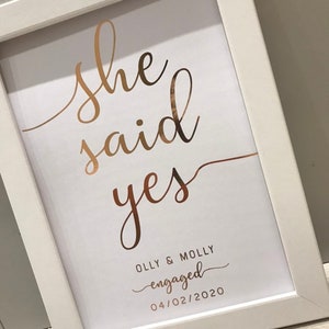 May include: A white framed print with the words "She said yes" in gold script. Below the text is the phrase "Olly & Molly engaged 04/02/2020" in a smaller font.