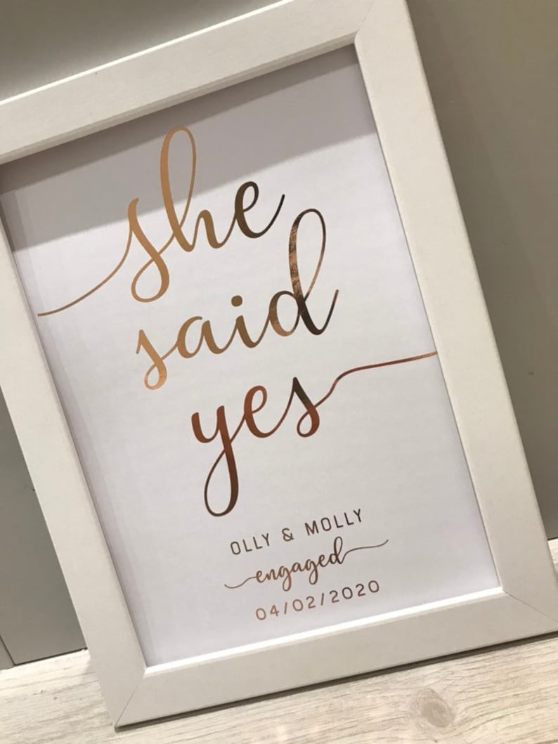 May include: A white framed print with the words "She said yes" in gold script. Below the text is the phrase "Olly & Molly engaged 04/02/2020" in a smaller font.