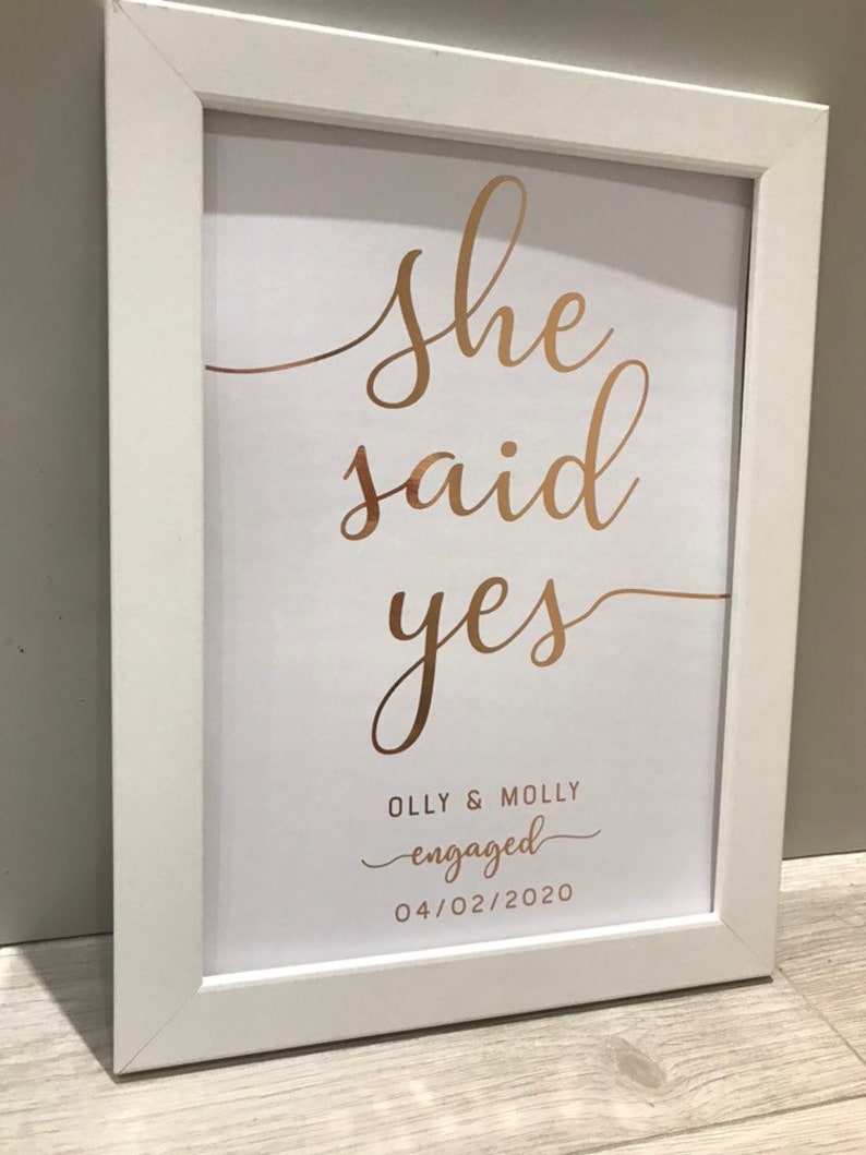 May include: White framed print with gold lettering that says "She said yes" with the names "Olly & Molly" and the text "engaged" underneath. The date "04/02/2020" is printed below the text.