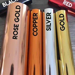 May include: Close-up of six rolls of metallic vinyl adhesive film in various colors: black, rose gold, copper, silver, gold, and red. The rolls are arranged horizontally, with the labels facing the viewer.