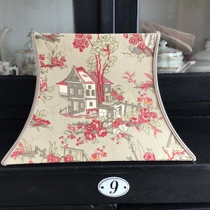 Puede incluir: Una pantalla de lámpara beige con un estampado floral y de casa en rojo, gris y crema. La pantalla tiene una forma curva y un ribete color crema. El diseño presenta una casa, árboles y flores. La pantalla está sobre una superficie negra.