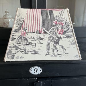 Op de afbeelding: Een trapeziumvormige lampenkap met een crèmekleurige achtergrond en een zwarte inktillustratie van een strandtafereel. Het ontwerp toont een rood-wit gestreepte tent, een persoon in een gestreept shirt en twee wandelende figuren op het zand.