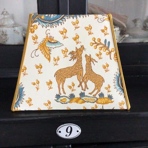 Puede incluir: Pantalla de lámpara decorativa con fondo crema y estampado de ciervos, pájaros y motivos florales en tonos dorados, azules y amarillos. La pantalla tiene forma trapezoidal y ribete dorado.