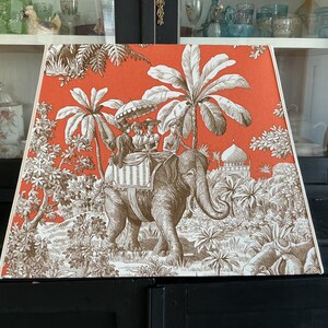 Può includere: Paralume trapezoidale con un motivo toile de Jouy marrone e arancione. L'illustrazione raffigura un elefante che trasporta persone, palme e un edificio. Lo sfondo è di colore arancione pieno.