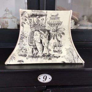Op de afbeelding: Crèmekleurige decoratieve plaat met een zwart-wit olifantenontwerp. De plaat heeft een gedetailleerde illustratie van een olifant die mensen, palmbomen en een pagode draagt. De plaat heeft een gebogen vorm en een lichtroze rand.