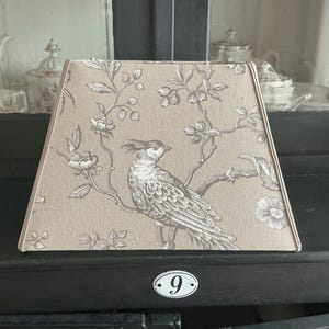 Puede incluir: Una pantalla de lámpara trapezoidal con base de tela color canela claro y un patrón estampado en marrón oscuro de un ave con cresta posada en una rama florida. La pantalla tiene un marco estructurado con bordes limpios y diseño botánico.