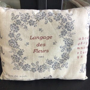 Könnte beinhalten: Beiges rechteckiges Kissen mit floralem Design und dem Text "Langage des Fleurs PARIS". Das florale Design ist ein Kranz aus grauen Blumen. Auf der Seite ist ein Alphabet aufgedruckt.