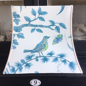 Könnte beinhalten: Quadratischer Lampenschirm mit blauem Vogel- und Schmetterlingsdesign auf weißem Hintergrund. Der Vogel sitzt auf einem Ast mit blauen Blättern. Der Lampenschirm hat eine cremefarbene Umrandung.