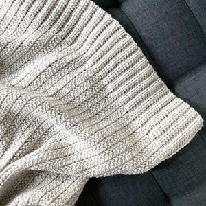May include: A close-up of a crocheted throw blanket in a light beige color. The blanket is made with a simple stitch pattern and has a soft, textured look.