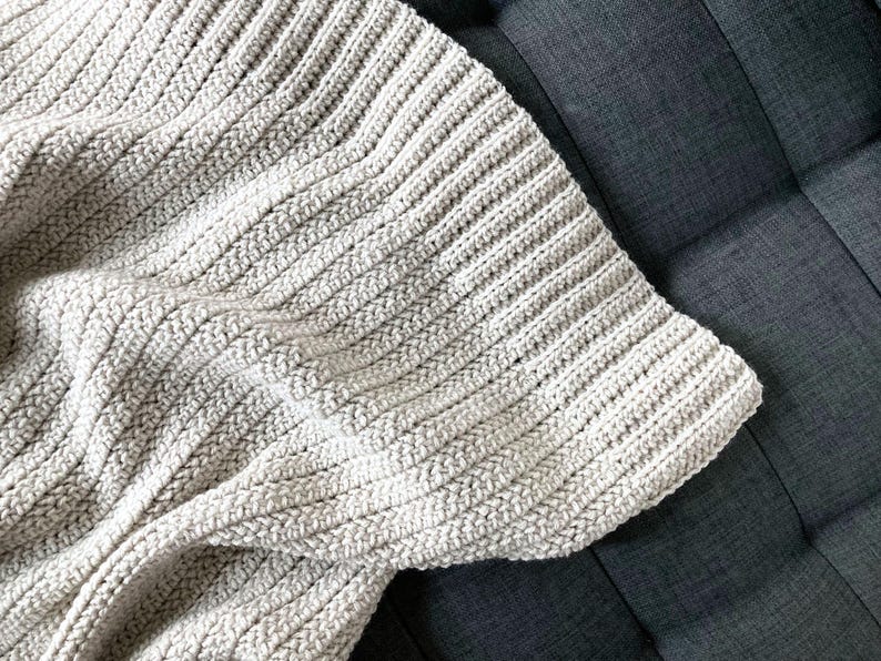 May include: A close-up of a crocheted throw blanket in a light beige color. The blanket is made with a simple stitch pattern and has a soft, textured look.