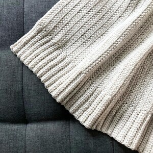 May include: A close-up of a crocheted throw blanket in a light beige color. The blanket is made with a simple stitch pattern and has a soft, textured feel.