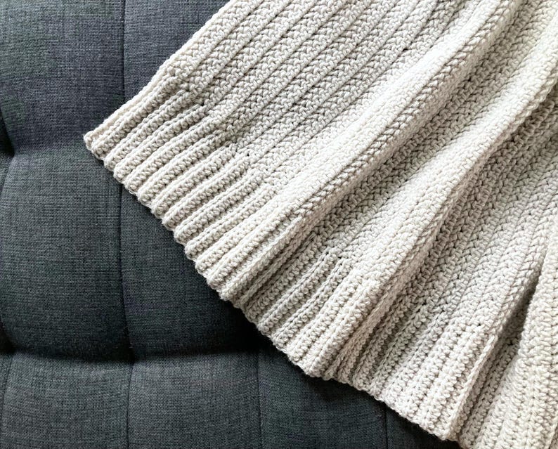 May include: A close-up of a crocheted throw blanket in a light beige color. The blanket is made with a simple stitch pattern and has a soft, textured feel.