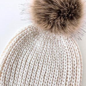 May include: Cream-colored knit beanie with a large, fluffy, brown faux fur pom-pom. The hat features a ribbed knit pattern and is designed for warmth and style. A cozy winter accessory.