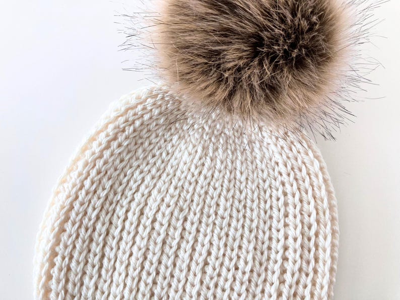 May include: Cream-colored knit beanie with a large, fluffy, brown faux fur pom-pom. The hat features a ribbed knit pattern and is designed for warmth and style. A cozy winter accessory.