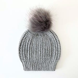 May include: A gray knit beanie with a large, fluffy, gray faux fur pom-pom on top. The hat has a ribbed knit design and is made of a soft, warm material. This winter hat is perfect for cold weather.