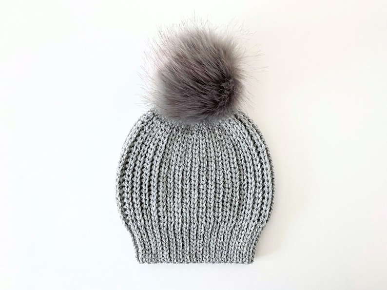 May include: A gray knit beanie with a large, fluffy, gray faux fur pom-pom on top. The hat has a ribbed knit design and is made of a soft, warm material. This winter hat is perfect for cold weather.