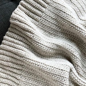 May include: A close-up of a soft, gray crocheted blanket with a textured, ribbed pattern. The blanket is folded and draped over a dark surface.
