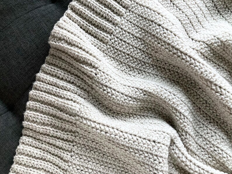 May include: A close-up of a soft, gray crocheted blanket with a textured, ribbed pattern. The blanket is folded and draped over a dark surface.