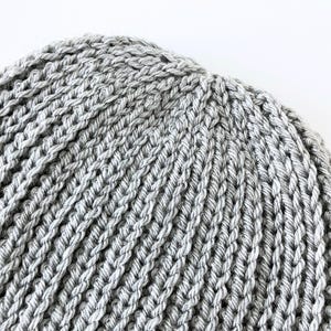 May include: Close-up of a gray knitted beanie hat. The hat is made of a thick, textured yarn with a ribbed pattern. The hat is a light gray color and appears to be handmade.