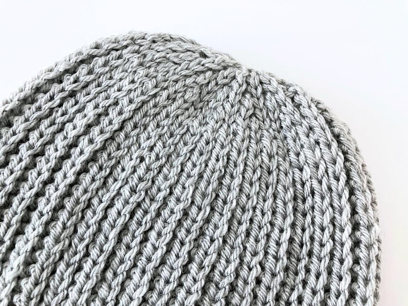 May include: Close-up of a gray knitted beanie hat. The hat is made of a thick, textured yarn with a ribbed pattern. The hat is a light gray color and appears to be handmade.