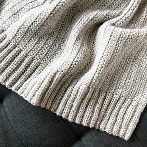 May include: A soft, beige crocheted throw blanket with a ribbed texture. The blanket is folded and draped over a gray couch.