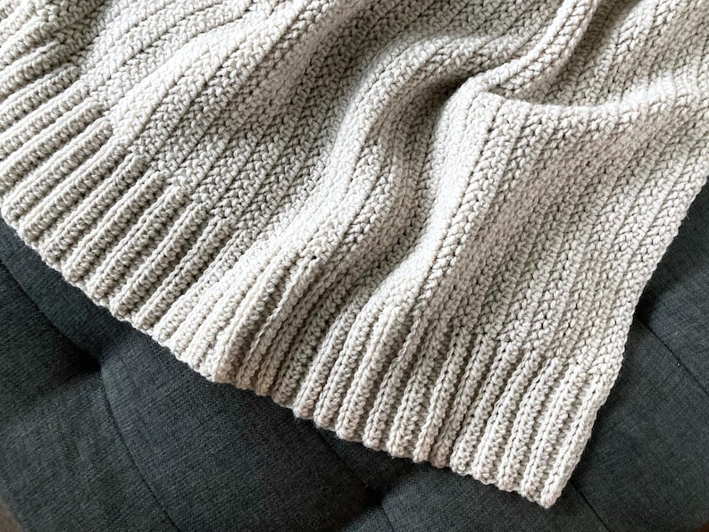 May include: A soft, beige crocheted throw blanket with a ribbed texture. The blanket is folded and draped over a gray couch.