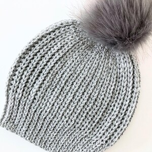 May include: A gray knitted beanie with a faux fur pom-pom. The hat is made of a thick, textured yarn and has a ribbed knit pattern. The pom-pom is a fluffy, light gray color.