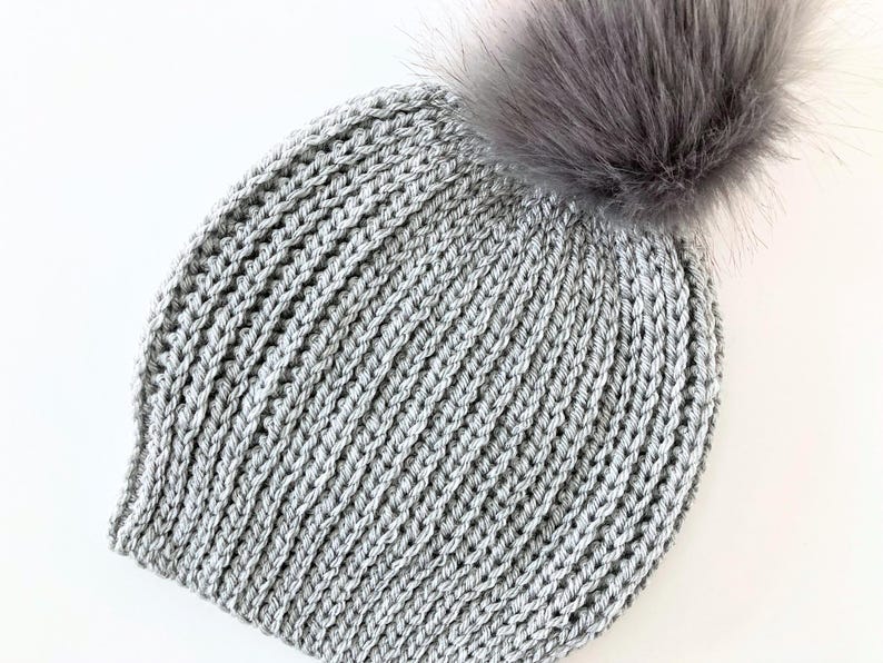 May include: A gray knitted beanie with a faux fur pom-pom. The hat is made of a thick, textured yarn and has a ribbed knit pattern. The pom-pom is a fluffy, light gray color.