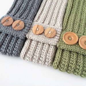 Puede incluir: Primer plano de tres calentadores de piernas tejidos en gris, beige y verde. Cada calentador de piernas tiene un diseño acanalado y está adornado con dos botones redondos de madera. Los calentadores de piernas se muestran sobre una superficie blanca.