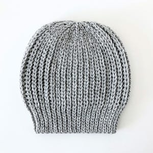 May include: A gray knitted beanie hat with a ribbed design. The hat is made of a soft, textured yarn and is suitable for cold weather. The hat is shown on a white background.