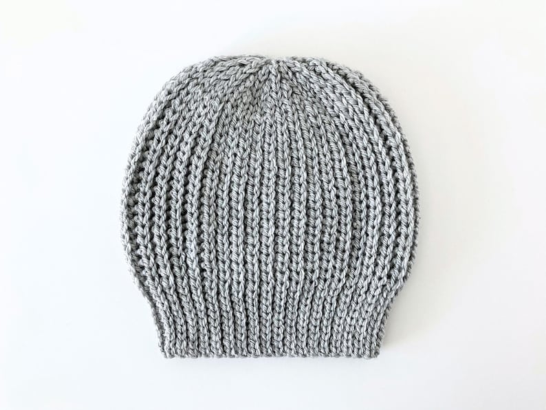 May include: A gray knitted beanie hat with a ribbed design. The hat is made of a soft, textured yarn and is suitable for cold weather. The hat is shown on a white background.