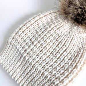 May include: A cream-colored knitted beanie with a fluffy brown faux fur pom pom on top.