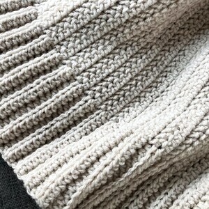 May include: Close-up of a beige crocheted blanket with a textured pattern. The blanket is made of soft yarn and has a warm, cozy feel.