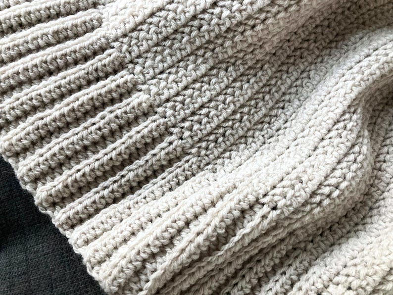 May include: Close-up of a beige crocheted blanket with a textured pattern. The blanket is made of soft yarn and has a warm, cozy feel.