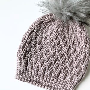 Puede incluir: Un gorro tejido de color malva claro con un patrón de diamantes y un pompón de piel sintética gris. Este accesorio de invierno está hecho de un material de punto suave.