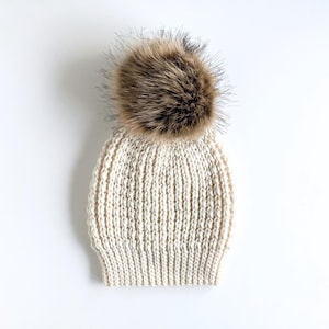 May include: A cream-colored knitted beanie with a brown faux fur pom-pom on top.