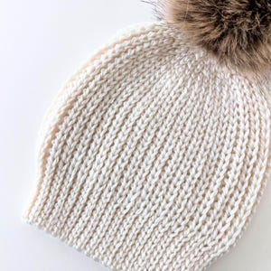 May include: Cream-colored knit hat with a large, fluffy brown pom-pom. The hat features a ribbed knit pattern and is made of a soft, warm material. A winter accessory for cold weather.