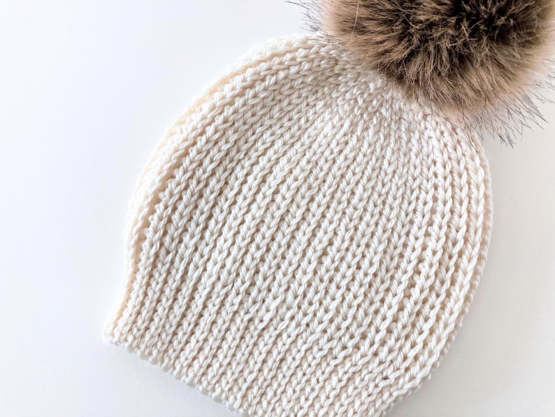 May include: Cream-colored knit hat with a large, fluffy brown pom-pom. The hat features a ribbed knit pattern and is made of a soft, warm material. A winter accessory for cold weather.