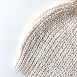 May include: Close-up of a cream-colored knitted beanie hat with a brown faux fur pom-pom. The hat features a ribbed knit design and is made of a soft, warm material, perfect for cold weather.