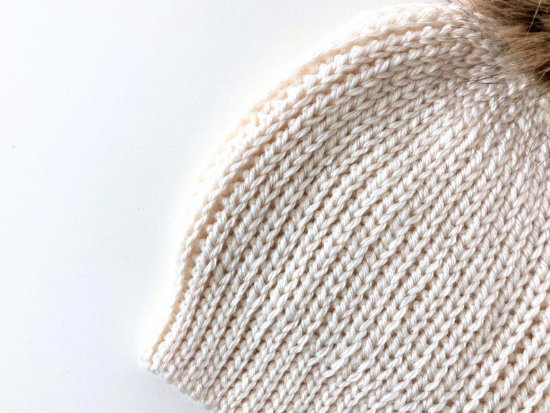 May include: Close-up of a cream-colored knitted beanie hat with a brown faux fur pom-pom. The hat features a ribbed knit design and is made of a soft, warm material, perfect for cold weather.
