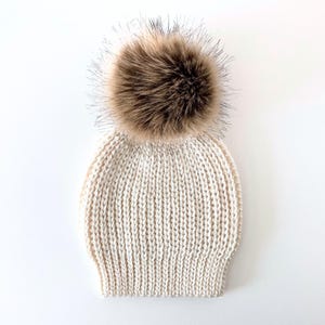 May include: Cream-colored knit beanie with a large, fluffy brown faux fur pom-pom. The hat features a ribbed knit design and is set against a white background. A winter accessory.
