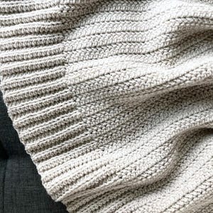 May include: A close-up of a soft, light gray crocheted blanket with a textured, ribbed pattern.