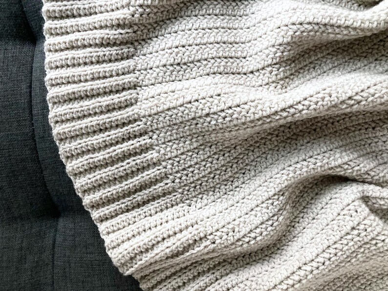 May include: A close-up of a soft, light gray crocheted blanket with a textured, ribbed pattern.
