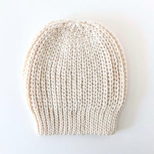 May include: Cream-colored knitted beanie hat. The hat is made of a thick, textured yarn with a ribbed design. The hat is a neutral color and would be suitable for any gender.