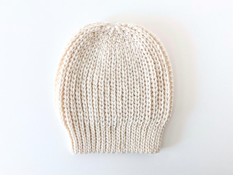 May include: Cream-colored knitted beanie hat. The hat is made of a thick, textured yarn with a ribbed design. The hat is a neutral color and would be suitable for any gender.