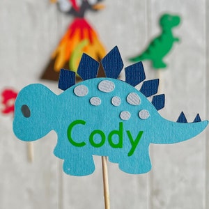 May include: Blue dinosaur cake topper with a green name "Cody" on the body. The dinosaur has a blue and white spiky back and is on a wooden stick.