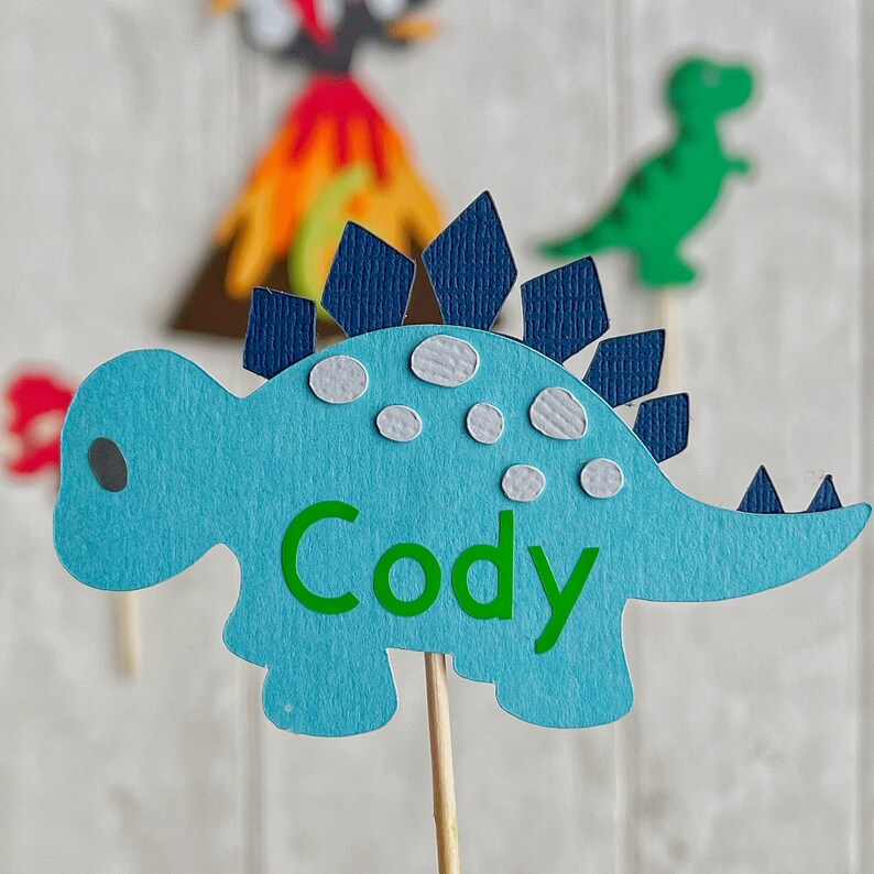 May include: Blue dinosaur cake topper with a green name "Cody" on the body. The dinosaur has a blue and white spiky back and is on a wooden stick.